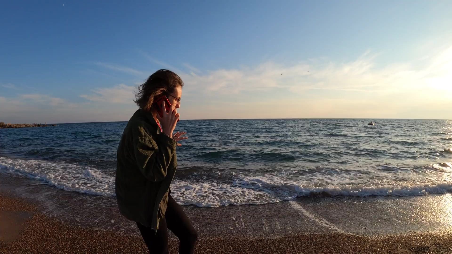 Una mujer camina por la orilla del mar al atardecer mientras habla por teléfono.